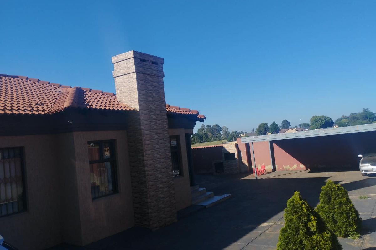 Rear exterior view of the property featuring a tiled roof, stone-clad chimney, paved driveway, carport, and a built-in braai area.