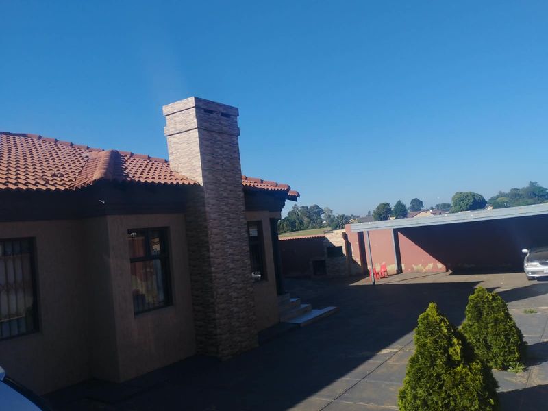 Rear exterior view of the property featuring a tiled roof, stone-clad chimney, paved driveway, carport, and a built-in braai area.