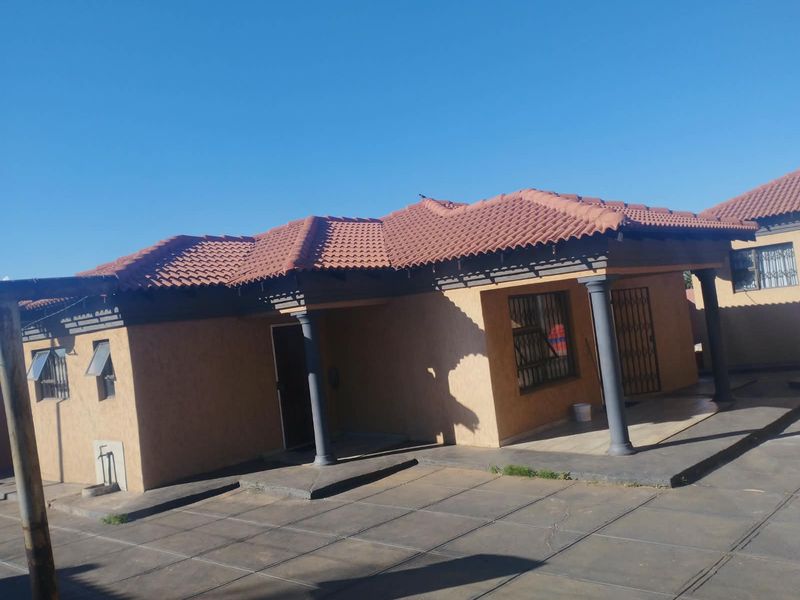 Front view of a single-story house featuring a terracotta tiled roof, textured facade, covered patio area with columns, and a paved driveway.