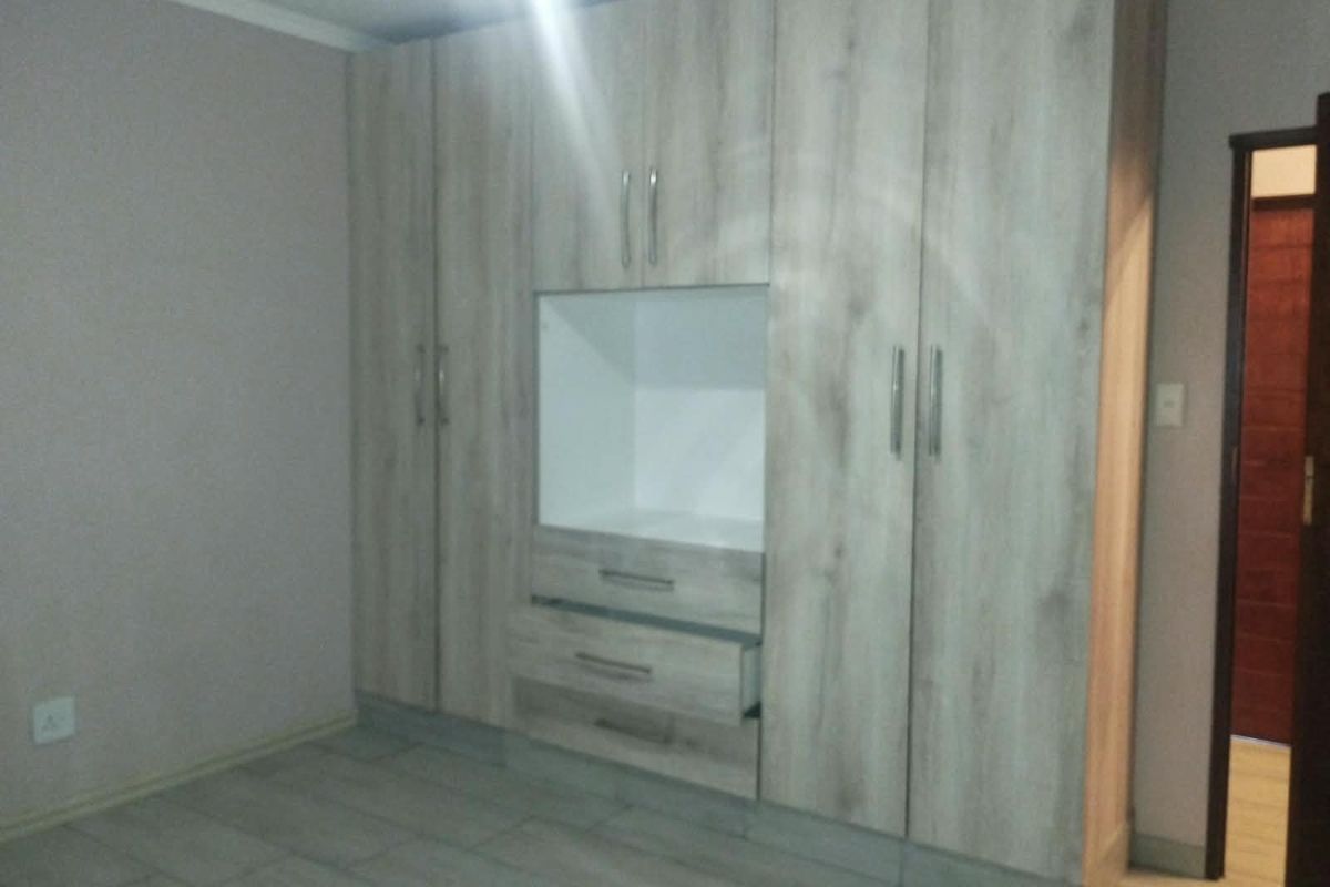 A clean bedroom featuring modern light wood-finish built-in cupboards with integrated drawers and neutral tiled flooring.