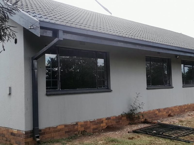 Rear elevation of a single-story house featuring a tiled roof, grey rendered walls, brick plinth, and metal-framed windows with security bars.