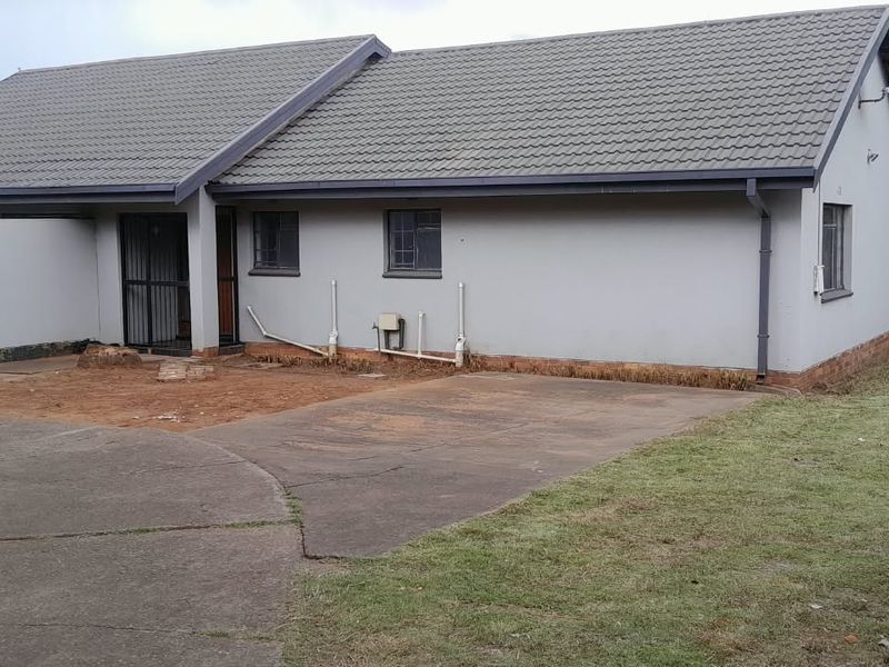 Rear view of the property featuring a concrete driveway, tiled roof, and surrounding garden area with visible exterior maintenance requirements.
