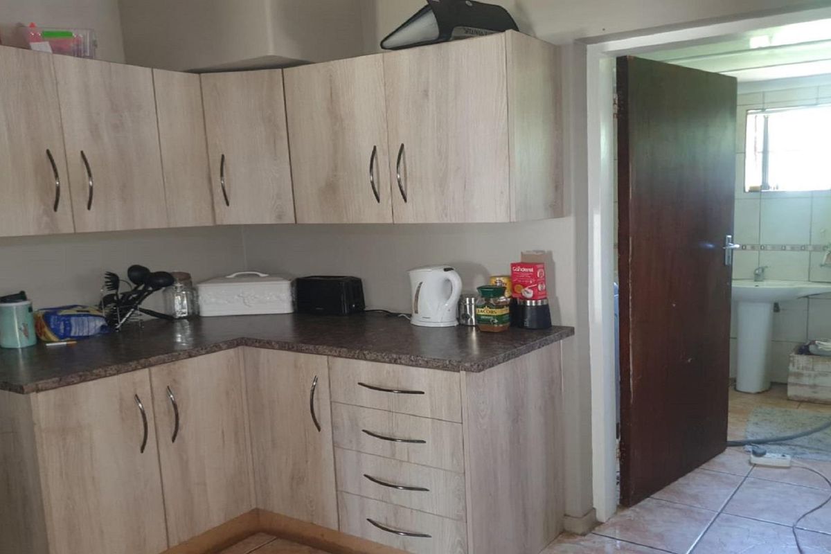 A functional kitchen featuring light wood-grain cabinetry, dark laminate countertops, and tiled flooring, with an adjacent view into a tiled bathroom.