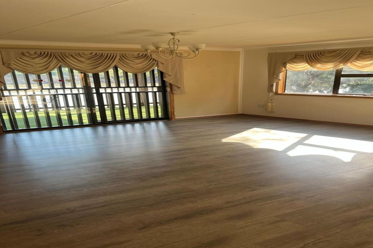 A spacious, sunlit living room featuring modern wood-look laminate flooring, large windows, and a sliding glass door with security gate access to the garden.