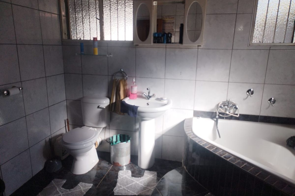 A dated bathroom featuring a corner bathtub, pedestal basin, and toilet with full-height wall tiling and security bars on the windows.