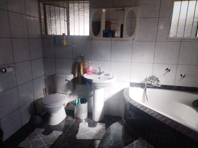 A dated bathroom featuring a corner bathtub, pedestal basin, and toilet with full-height wall tiling and security bars on the windows.