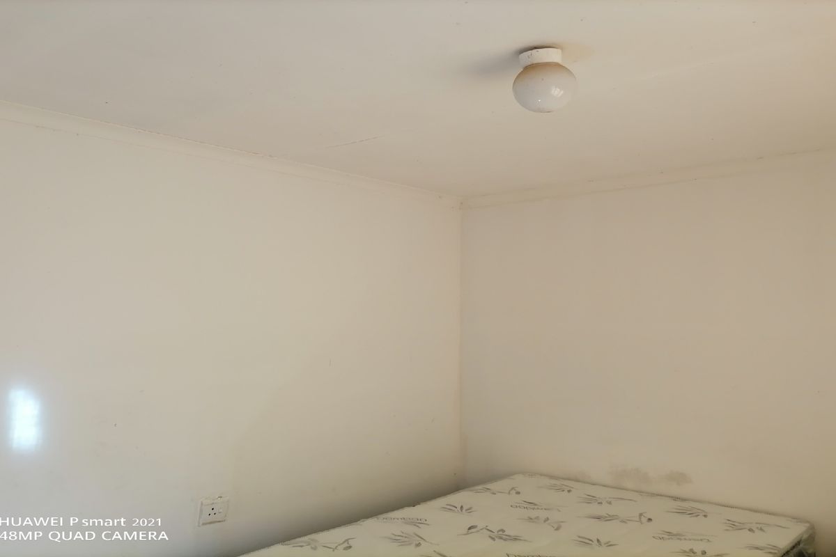 A simple, empty bedroom space featuring plain white walls, a ceiling-mounted light fixture, and a single electrical outlet.