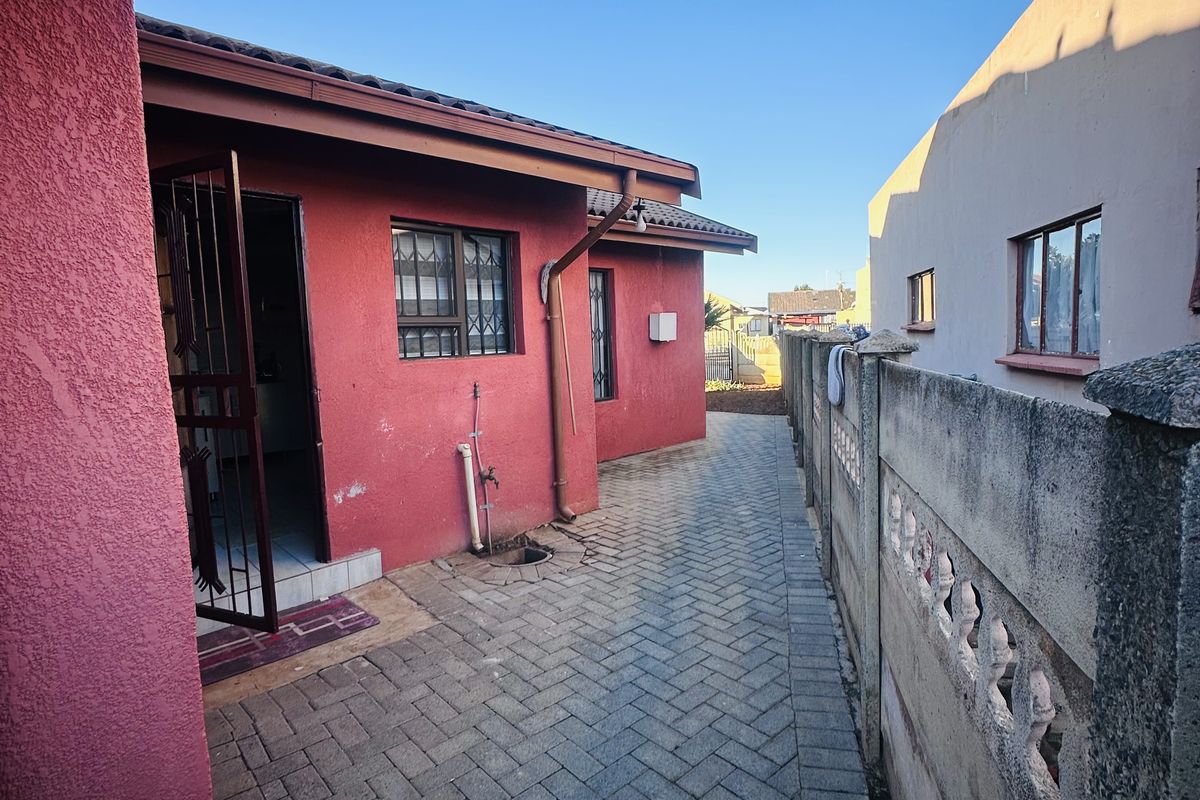 A side exterior view of the property featuring paved walkways, a red textured facade, security gates, and a concrete boundary wall.