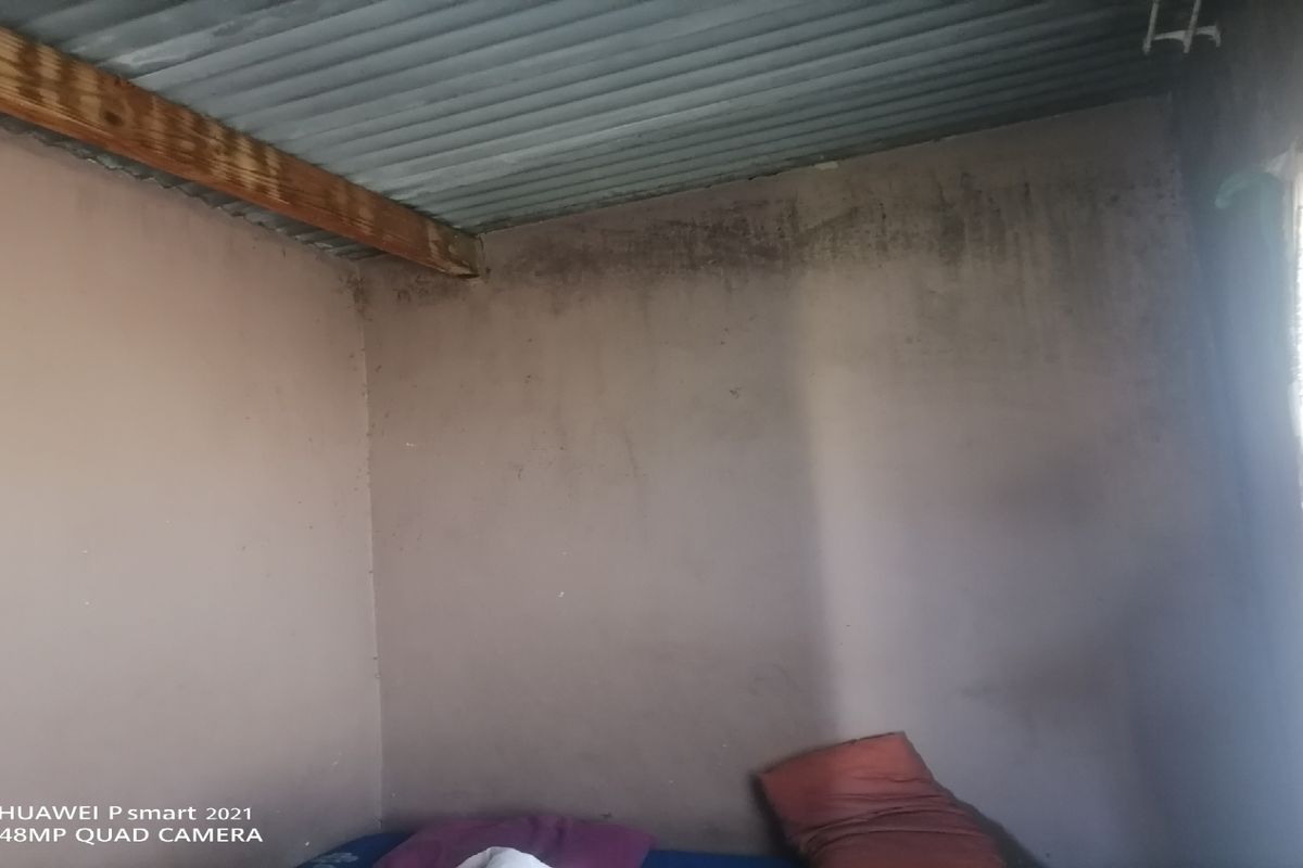 A basic bedroom space featuring exposed corrugated metal ceiling and unfinished wall surfaces showing signs of wear and staining.