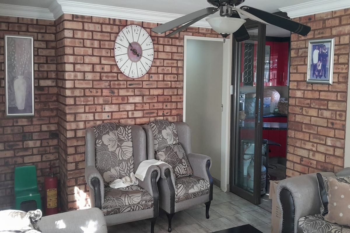 Living area featuring a prominent exposed brick feature wall, tiled flooring, and a ceiling fan, with an adjacent view into the kitchen area.