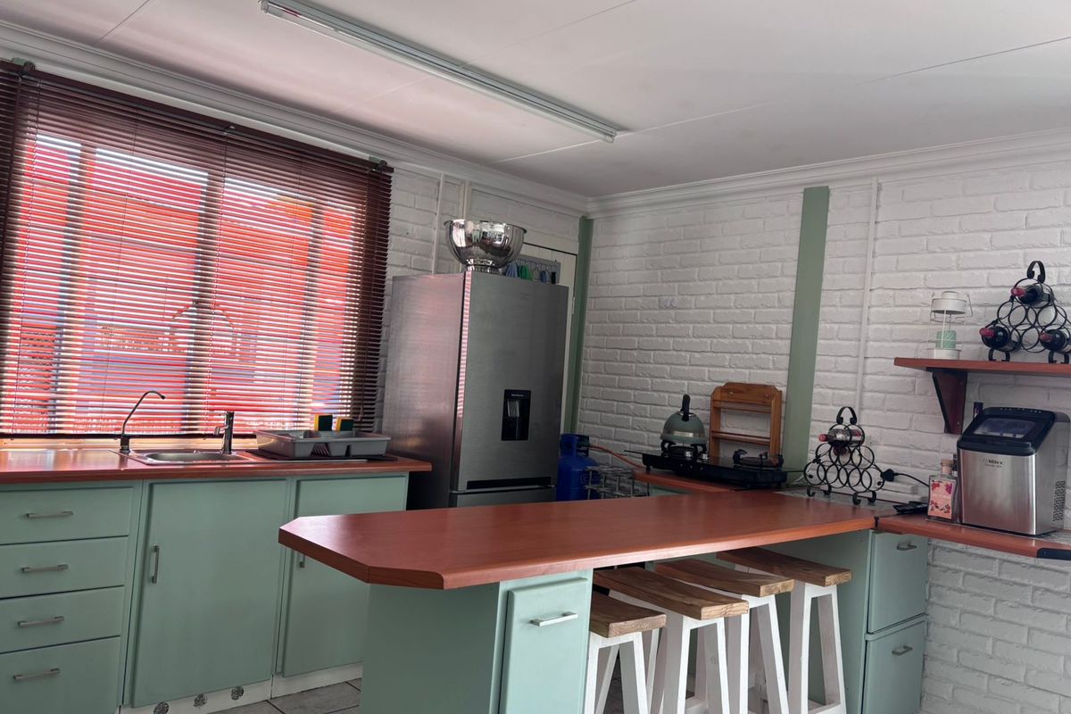 A functional kitchen featuring light green cabinetry, a wooden breakfast bar, tiled flooring, and a large window with venetian blinds.