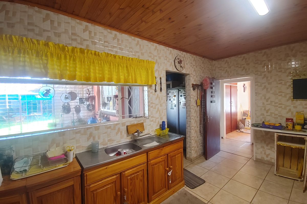 A dated kitchen featuring wooden cabinetry, a stainless steel sink, tiled walls, and a timber-paneled ceiling, with an adjacent doorway leading to another room.