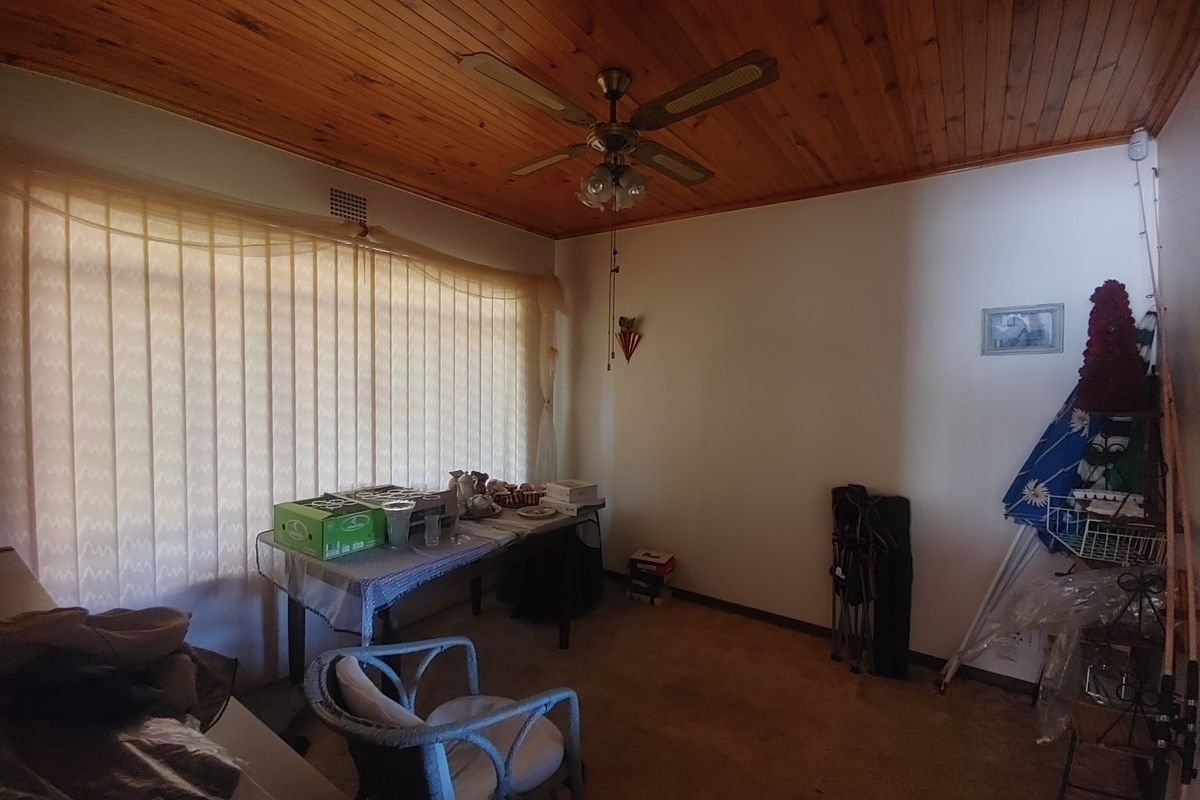 A living room featuring a timber-paneled ceiling, ceiling fan, and large window with vertical blinds, offering a dated interior space ready for modernization.
