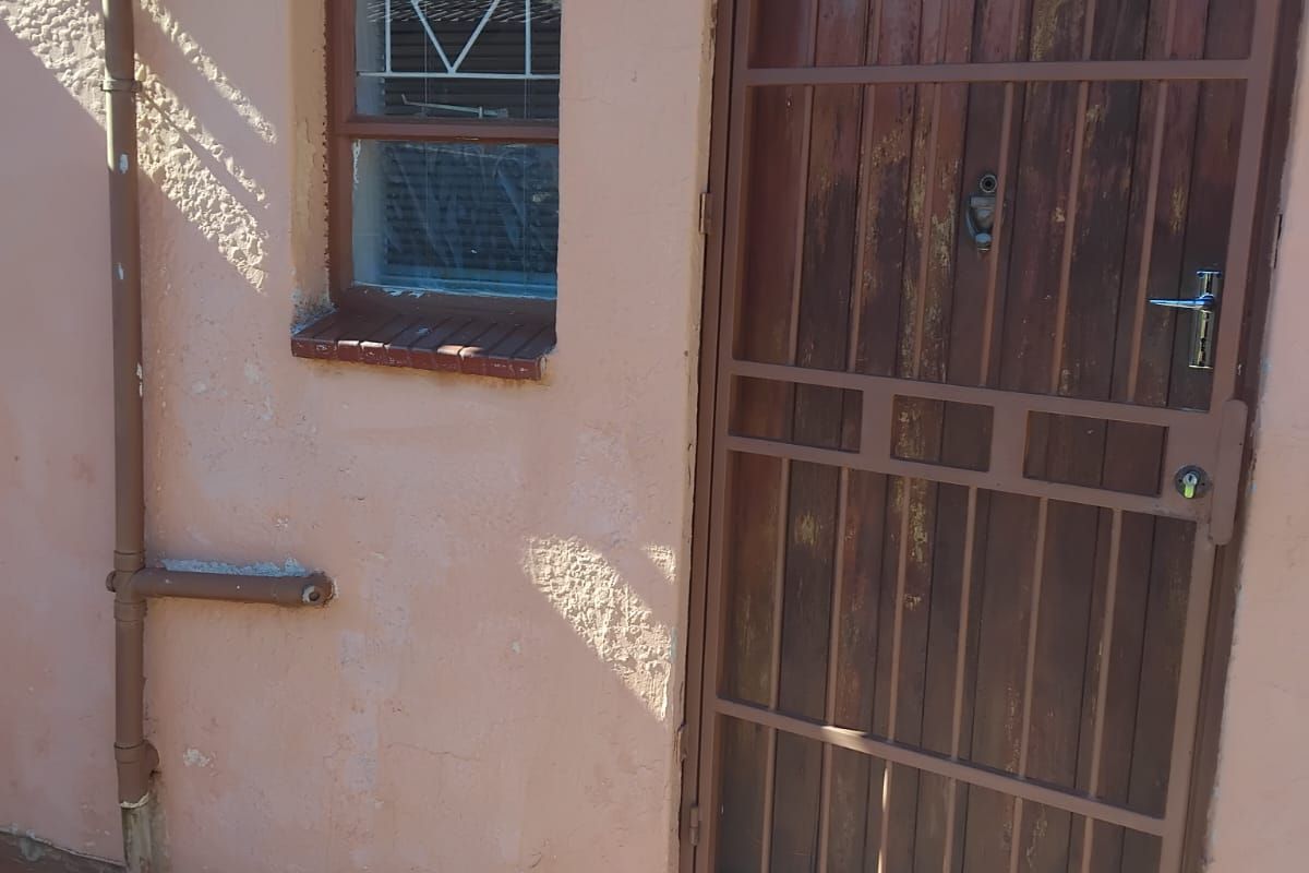 Exterior view of a rear entrance featuring a timber door with a security gate, adjacent window, and paved ground area.