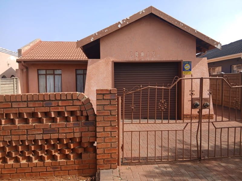 Front view of a single-story house featuring a garage, paved driveway, and brick boundary wall with a metal security gate.