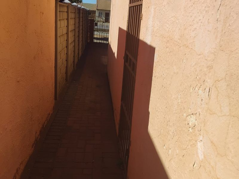 A narrow side passage featuring paved flooring, a security gate, and boundary walling, showing signs of wear on the exterior plaster.
