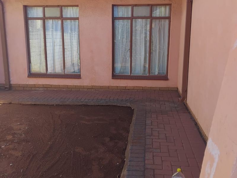 Rear exterior view of the house featuring brick paving, tiled roof, and security-barred windows, with an unfinished garden area.