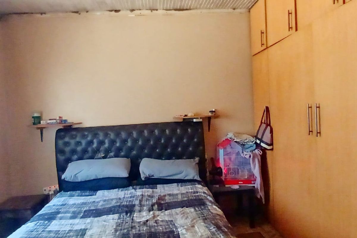 A bedroom featuring built-in wooden cupboards and a window with curtains. The ceiling shows visible signs of water damage.