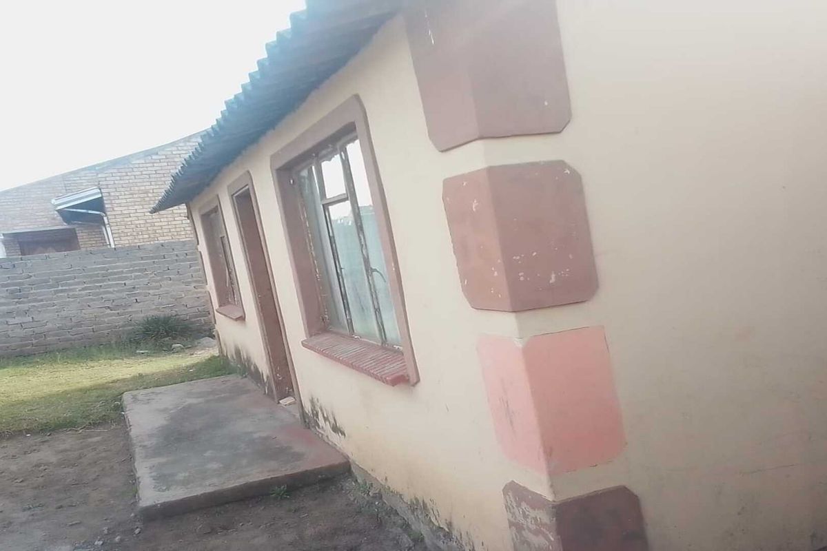 Rear exterior view of a single-story house featuring a concrete patio, metal window frames, and visible signs of exterior wall weathering and damp.