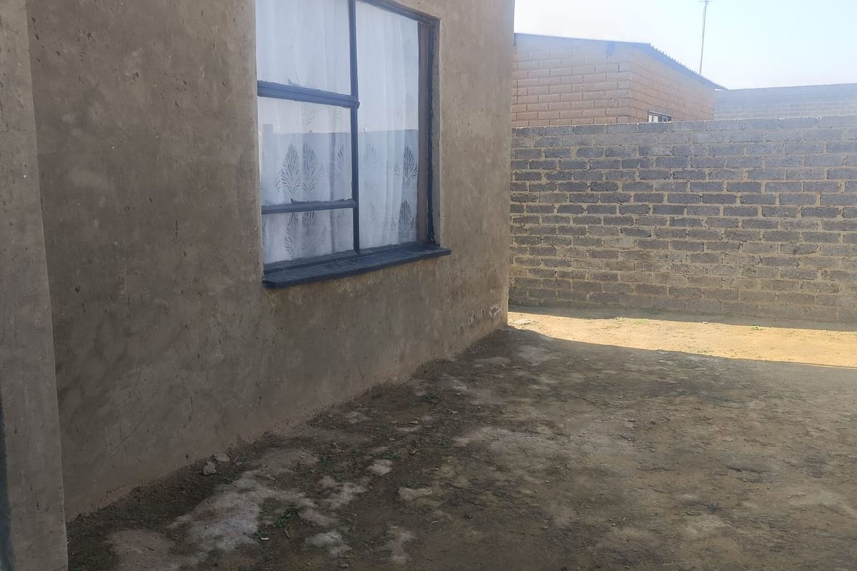 Exterior view of the property featuring an unfinished plastered wall, a metal-framed window, and an unpaved yard area enclosed by a brick boundary wall.