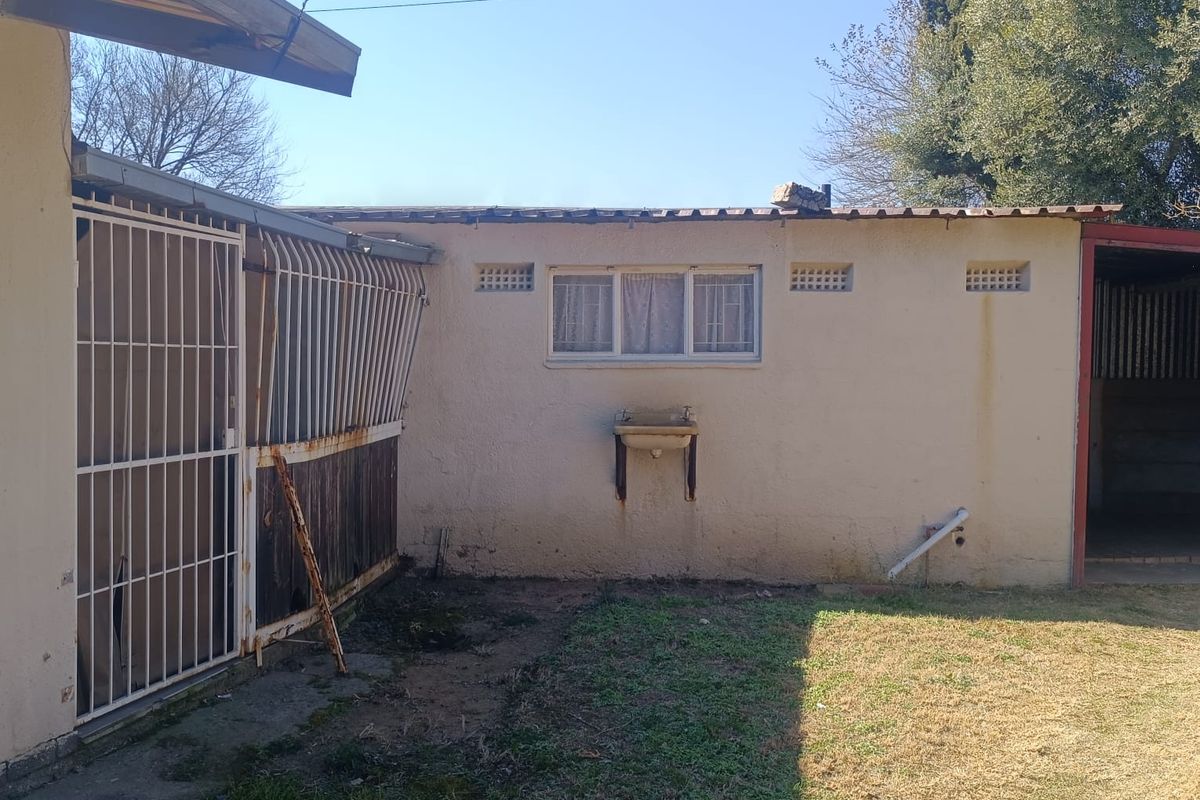 Rear exterior view of the property featuring a security gate, wall-mounted basin, and basic garden area with visible signs of wear and maintenance needs.