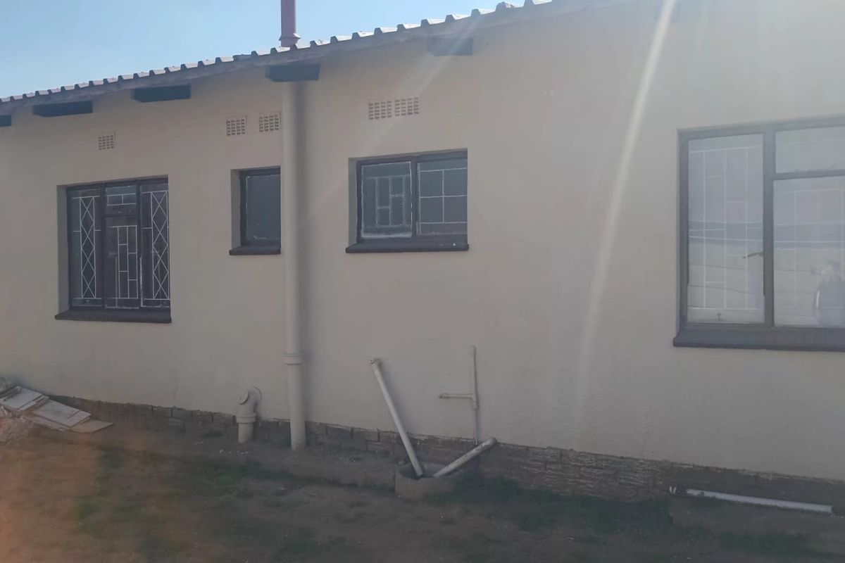 Rear exterior view of the house featuring metal window frames with security bars, corrugated roofing, and exposed plumbing infrastructure along the wall.