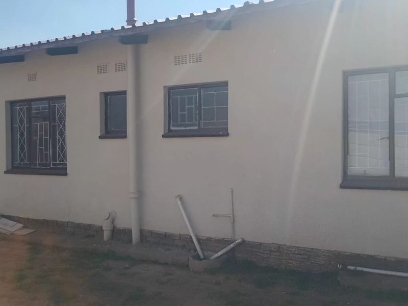 Rear exterior view of the house featuring metal window frames with security bars, corrugated roofing, and exposed plumbing infrastructure along the wall.