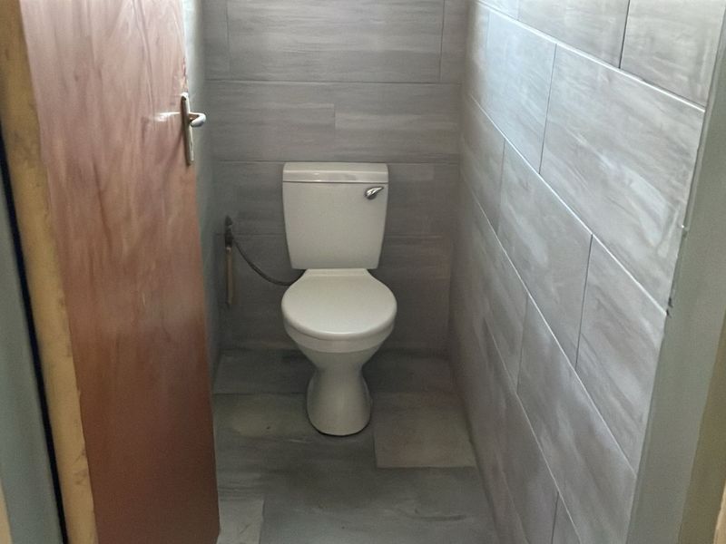 A compact bathroom featuring a standard toilet, grey tiled walls, and tiled flooring, currently requiring minor finishing touches to the door frame.
