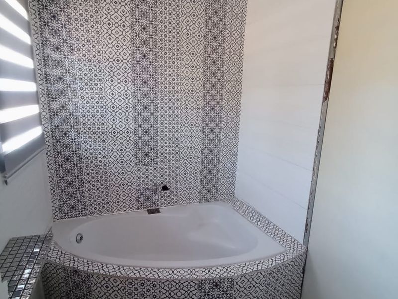 A bathroom featuring a corner bathtub with decorative patterned tiling on the walls and floor, complemented by a modern roller blind.