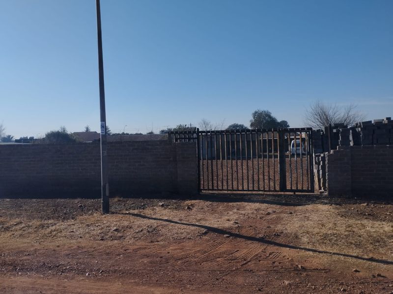 Front view of the property featuring a brick boundary wall and a metal driveway gate, with an unpaved approach.