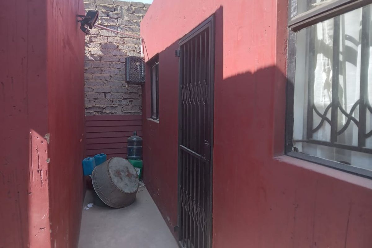 A narrow exterior side passage featuring red painted walls, security gates, and concrete paving, showing signs of wear and exposed brickwork.
