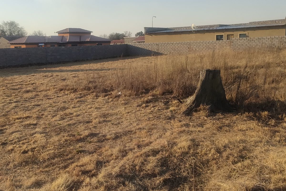 A vacant residential plot featuring dry grass, a tree stump, and perimeter boundary walls, situated in a developed residential neighborhood.