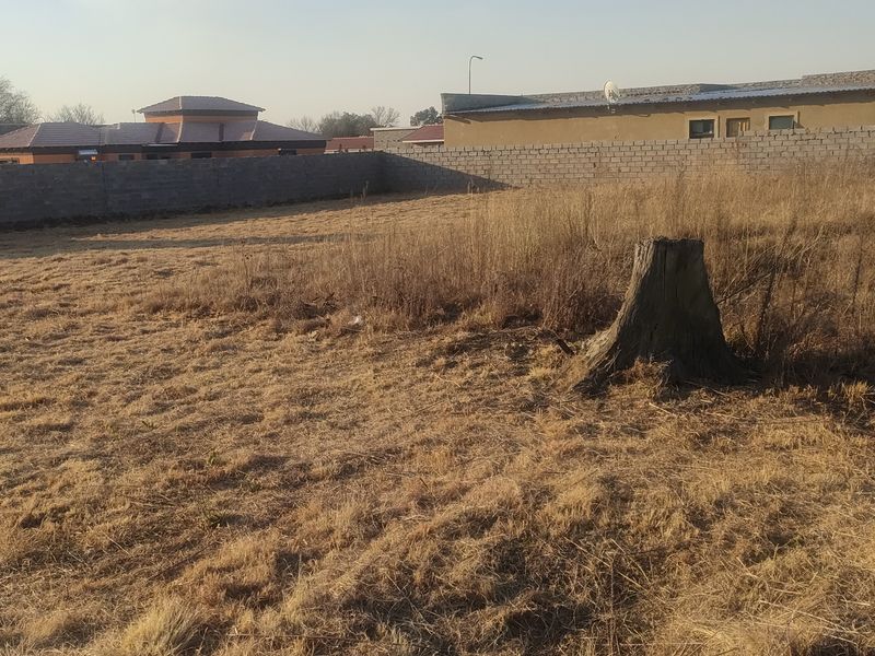 A vacant residential plot featuring dry grass, a tree stump, and perimeter boundary walls, situated in a developed residential neighborhood.