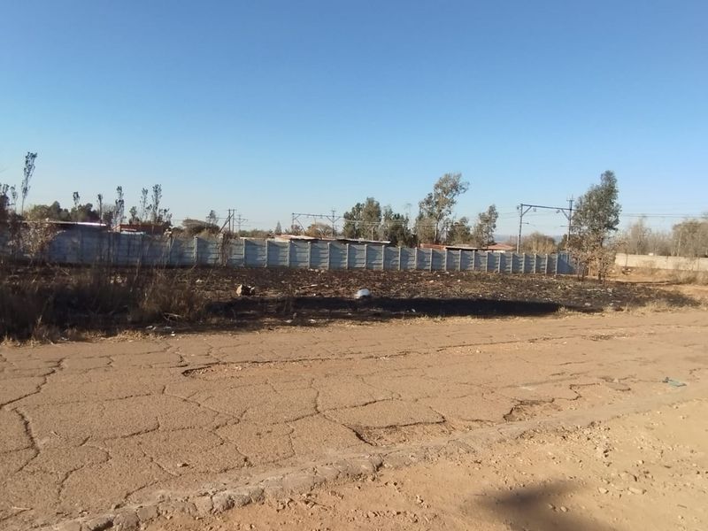 A vacant residential plot featuring a precast concrete boundary wall, accessible via an unpaved road with visible surface cracking.
