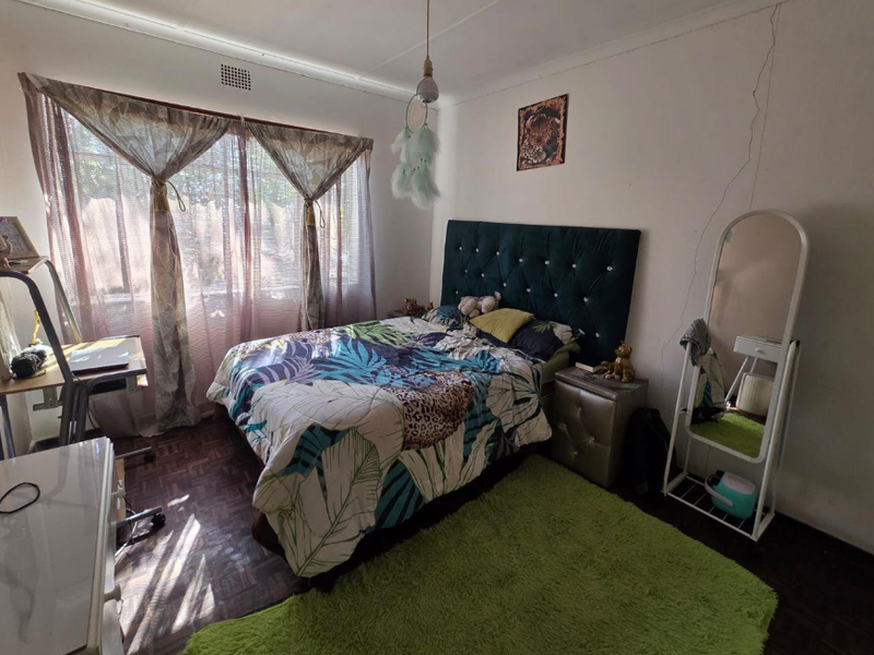 A bedroom featuring parquet flooring and a large window, currently showing signs of wear including a visible wall crack.
