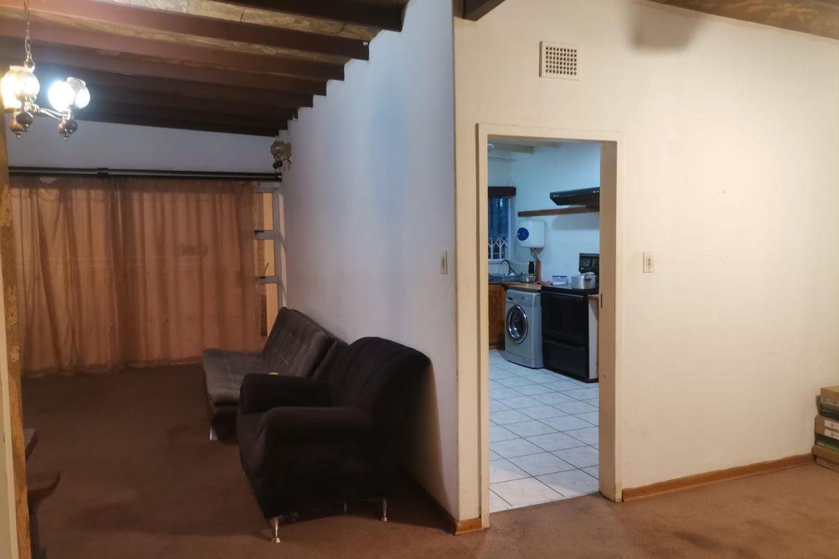 Living area featuring exposed timber ceiling beams and carpeted flooring, with an adjacent kitchen visible through the doorway.
