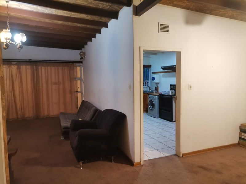 Living area featuring exposed timber ceiling beams and carpeted flooring, with an adjacent kitchen visible through the doorway.