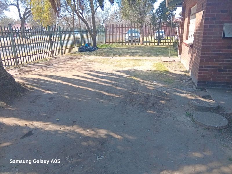 Front exterior view of the property featuring a brick facade, metal perimeter fencing, and a concrete driveway area requiring landscaping and maintenance.