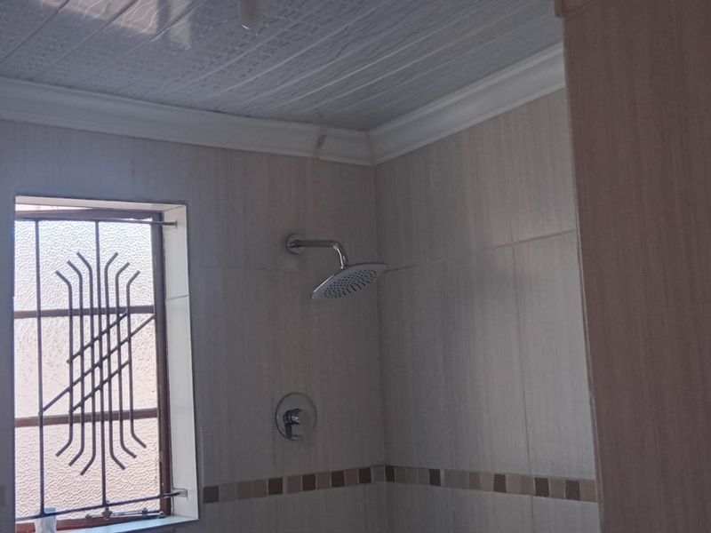 A functional bathroom shower area featuring tiled walls, a modern shower head, and security bars on the window.