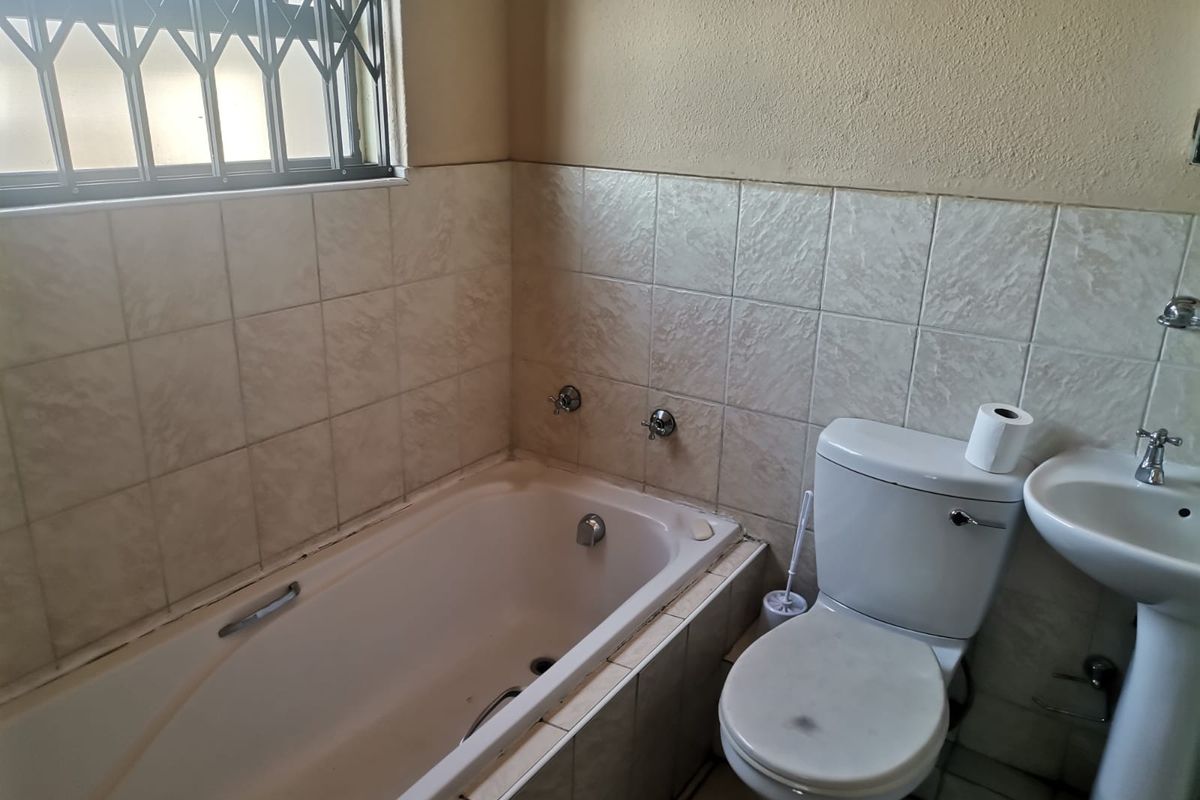 A dated bathroom featuring a built-in bathtub, pedestal basin, and toilet, with tiled walls and a security gate on the window.