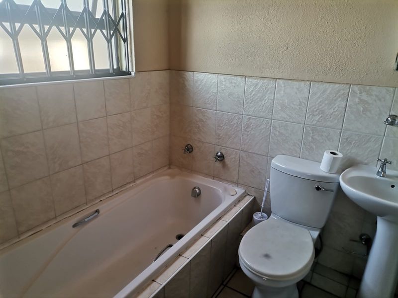 A dated bathroom featuring a built-in bathtub, pedestal basin, and toilet, with tiled walls and a security gate on the window.