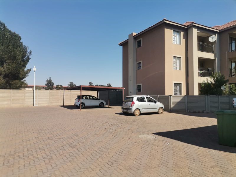 Exterior view of an apartment complex featuring a paved parking area with a covered carport and perimeter boundary walling.