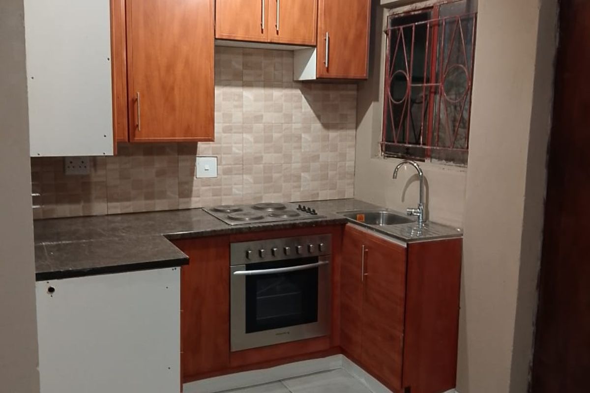 Compact kitchen featuring wood-finish cabinetry, built-in oven and hob, tiled backsplash, and neutral floor tiles. Requires minor finishing touches to cabinetry and walls.