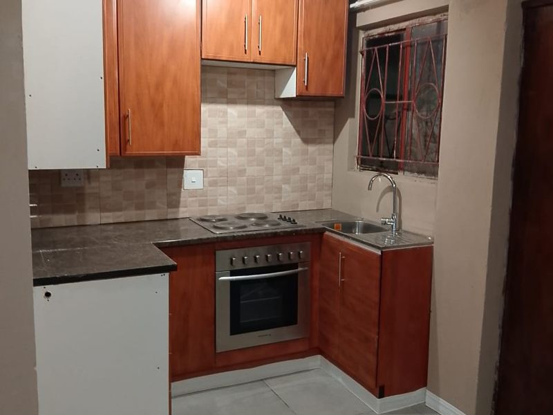 Compact kitchen featuring wood-finish cabinetry, built-in oven and hob, tiled backsplash, and neutral floor tiles. Requires minor finishing touches to cabinetry and walls.