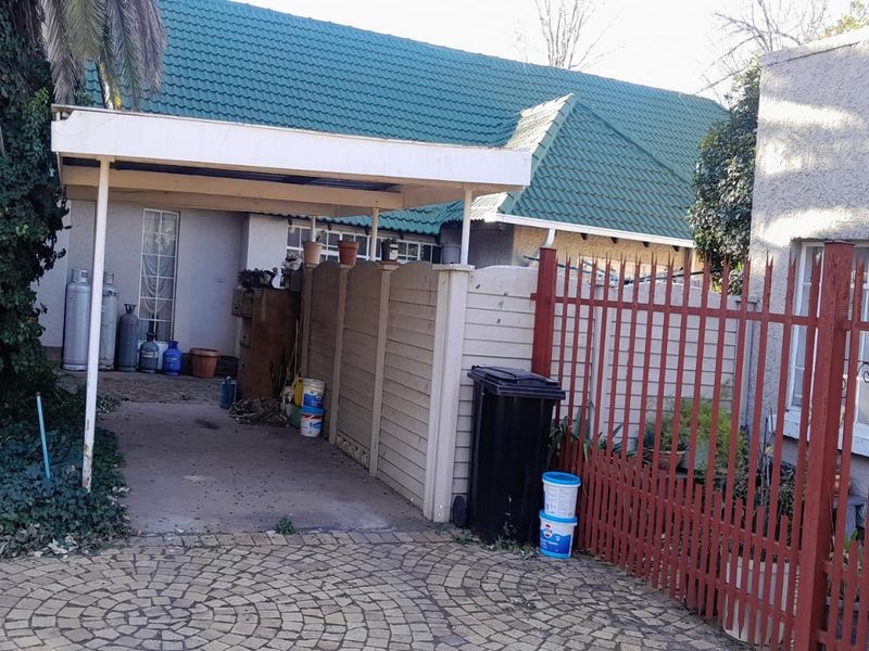 Front exterior view showing a paved driveway, carport structure, and metal security fencing with a tiled roof house facade.