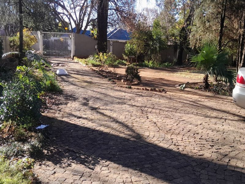 A front exterior view featuring a cobblestone driveway leading to a metal security gate, surrounded by mature trees and established garden landscaping.