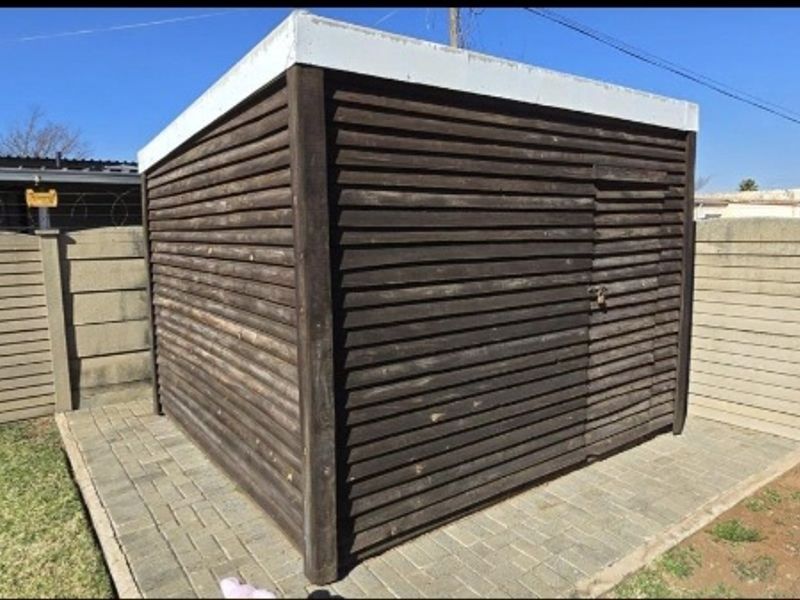 A detached timber-clad utility shed or storage structure situated on a paved base within a fenced garden area.