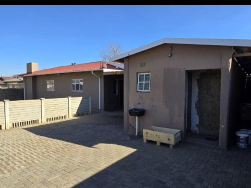 Rear exterior view showing a paved driveway, precast boundary wall, carport, and an external wash basin with visible wall plaster damage.