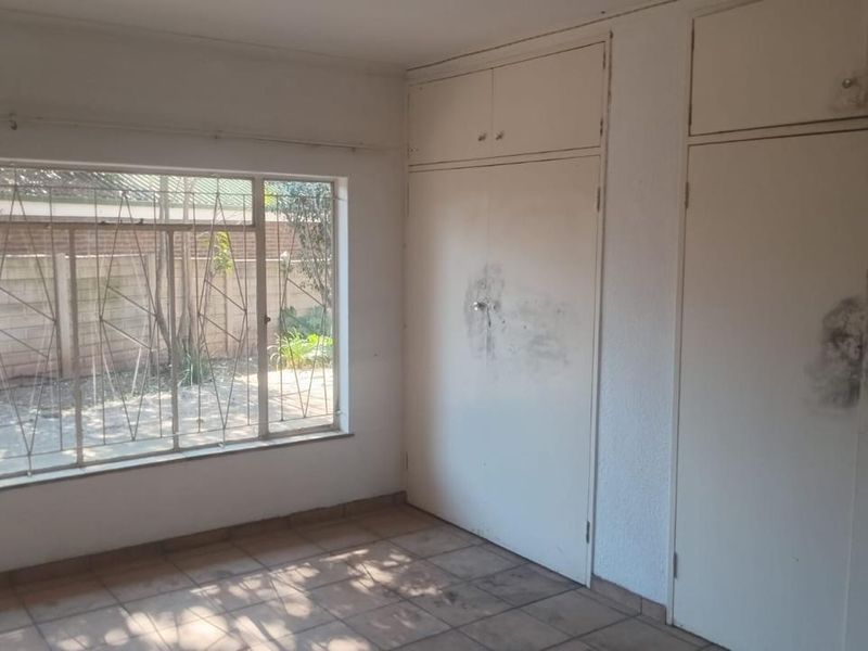 An empty bedroom featuring built-in cupboards, tiled flooring, and large windows with security bars, requiring cosmetic updates and maintenance.