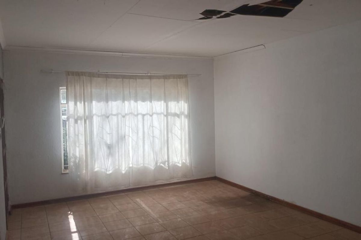 An empty living room featuring tiled flooring and a large window, currently requiring ceiling repairs.